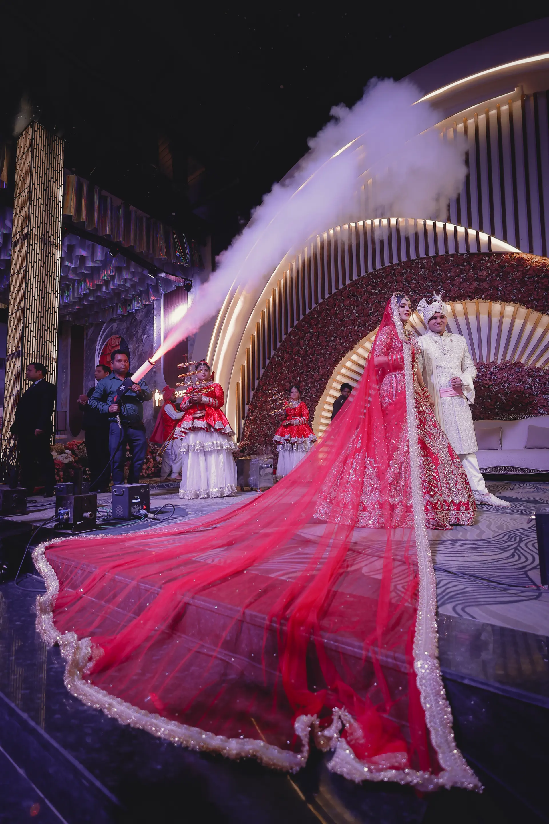 Grand stage entry of Riya & Akshay under smoke cannons with a long red veil train by The Wedding Galaxy